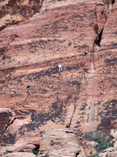 Three people are rock climbing on a steep, rugged red rock surface. The rock face has a textured appearance with a mix of red and black hues. The climbers are wearing harnesses and appear to be mid-climb, relying on the natural crevices and formations for support.