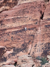 Three people are rock climbing on a steep, rugged red rock surface. The rock face has a textured appearance with a mix of red and black hues. The climbers are wearing harnesses and appear to be mid-climb, relying on the natural crevices and formations for support.