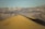 A vast desert landscape with towering sand dunes in the foreground and rugged mountains in the background. Several people are seen walking along the ridge of the sand dunes, creating a sense of adventure and exploration. The lighting suggests a late afternoon or early morning setting.