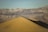 A vast desert landscape with towering sand dunes in the foreground and rugged mountains in the background. Several people are seen walking along the ridge of the sand dunes, creating a sense of adventure and exploration. The lighting suggests a late afternoon or early morning setting.