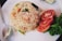 A plate features a serving of fried rice with visible vegetables such as diced carrots and leafy greens. Next to the rice are slices of fresh tomato and a garnish of green lettuce. A metal spoon is placed beside the dish, and a wedge of lime is partially visible in the corner.