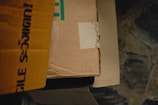 Cardboard boxes are stacked with a focus on one sealed with tape. The lighting is dim, casting shadows around and emphasizing textures.