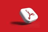 A three-dimensional square button with rounded edges featuring a red symbol resembling a ribbon on a white background. The button casts a shadow on a solid red background, suggesting a floating effect.