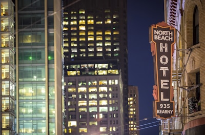 A cityscape at night featuring tall buildings with numerous illuminated windows. A prominent vintage-looking hotel sign with the text 'NORTH BEACH HOTEL 935 KEARNY' is visible on the right side, attached to a brick building. The modern glass skyscraper in the background contrasts with the older architectural style of the hotel.