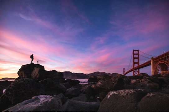 A breathtaking sunset with vibrant pink and purple hues fills the sky, casting a surreal light over a rocky shoreline. A person stands atop a large rock with a camera on a tripod, capturing the stunning view. In the background, the iconic structure of a large suspension bridge stretches across the scene, partially obscured by the rocky terrain.
