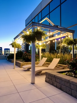 A modern rooftop terrace during twilight, featuring a sleek pergola adorned with string lights and hanging plants. Two stylish lounge chairs are positioned on a tiled floor surrounded by planters with lush green plants and flowers. The glass fa&ccedil;ade of the building reflects the evening sky, enhancing the urban ambiance.