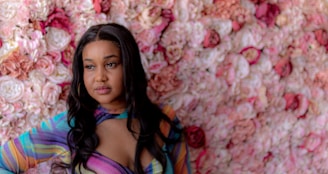 A woman stands in front of a vibrant wall of flowers with various shades of pink and red. She is wearing a colorful, striped top with an opening at the chest area. Her long, dark hair is styled with a middle part. The floral background creates a soft and romantic atmosphere.