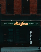 A storefront with a dark exterior and a neon sign displaying 'Miss Jones'. The words 'Cannabis' and 'Outposts' are illuminated in smaller letters above and below the name. To the side, a vertical 'OPEN' sign glows, inviting passersby. The building features brickwork and large, dark windows.