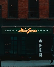 A storefront with a dark exterior and a neon sign displaying 'Miss Jones'. The words 'Cannabis' and 'Outposts' are illuminated in smaller letters above and below the name. To the side, a vertical 'OPEN' sign glows, inviting passersby. The building features brickwork and large, dark windows.