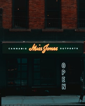 A storefront with a dark exterior and a neon sign displaying 'Miss Jones'. The words 'Cannabis' and 'Outposts' are illuminated in smaller letters above and below the name. To the side, a vertical 'OPEN' sign glows, inviting passersby. The building features brickwork and large, dark windows.