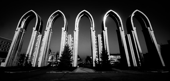 A series of large, modern arched structures made of light-colored material stand prominently against a dark sky. These arches feature an architectural style that emphasizes elongated, narrow forms. In front of the arches, several small coniferous trees are visible, suggesting a landscaped area. The background includes some buildings, hinting at an urban setting.