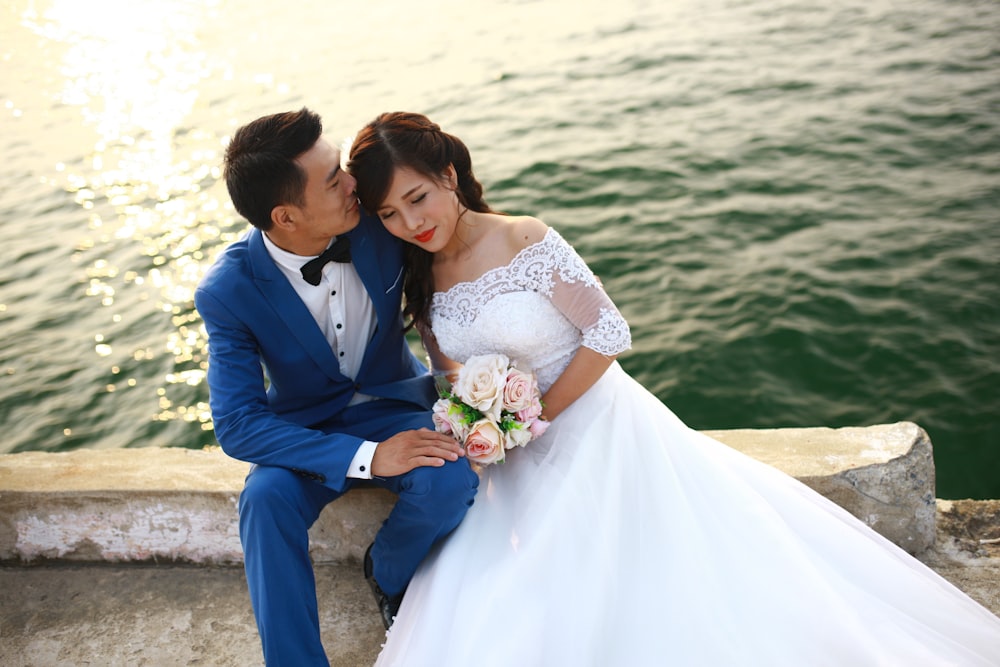 a man and woman sitting on a rock with a body of water behind them