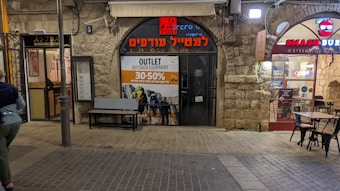 A street scene features storefronts with signage on a stone building. The main shop displays a sign for an outdoor equipment outlet sale offering discounts between 30 to 50 percent. Next door, a brightly lit fast-food restaurant called Smash Burger is visible. A person is walking on the left side of the frame, and a streetlamp is positioned centrally.