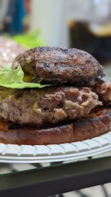 A close-up view of a hamburger with a portobello mushroom cap, a beef patty, and a leaf of lettuce, all placed on a toasted bun. The burger is on a white paper plate, and the background is blurred.