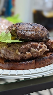 A close-up view of a hamburger with a portobello mushroom cap, a beef patty, and a leaf of lettuce, all placed on a toasted bun. The burger is on a white paper plate, and the background is blurred.