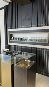 A display area features a sleek, modern design with dark vertical panels and a central shelf showcasing an array of small bottles. Two glass-topped pedestals beneath the shelf contain various textured pieces, possibly herbs or similar materials.