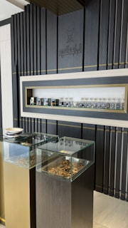 A display area features a sleek, modern design with dark vertical panels and a central shelf showcasing an array of small bottles. Two glass-topped pedestals beneath the shelf contain various textured pieces, possibly herbs or similar materials.