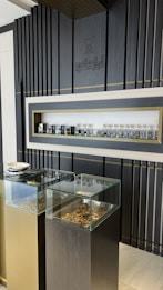 A display area features a sleek, modern design with dark vertical panels and a central shelf showcasing an array of small bottles. Two glass-topped pedestals beneath the shelf contain various textured pieces, possibly herbs or similar materials.