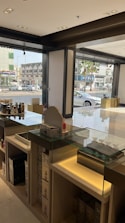 A well-lit modern store interior with glass display counters containing various products. The store has large windows offering a view of the busy street and nearby buildings. The shelves hold multiple boxes and items, and the setup suggests it’s a retail environment.