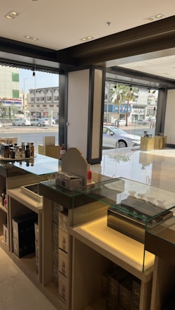A well-lit modern store interior with glass display counters containing various products. The store has large windows offering a view of the busy street and nearby buildings. The shelves hold multiple boxes and items, and the setup suggests it’s a retail environment.