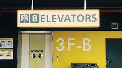 A bright yellow and black sign indicating the location of elevators, with prominent text and symbols. The background features yellow walls, with a number and letter in large print. Several smaller signs and a door are also visible, contributing to the organized atmosphere of a building's interior.