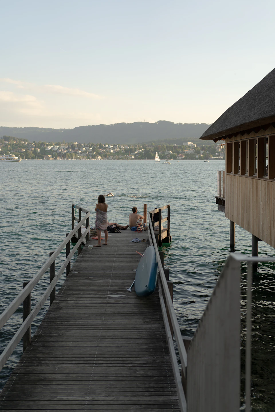 Two people stand on a wooden pier, one holding a surfboard, by a serene lake