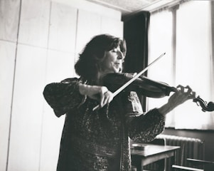 A person plays the violin in a softly lit room. The individual appears focused, wearing a patterned, long-sleeve outfit. Sunlight filters through a window with sheer curtains, creating a serene atmosphere.