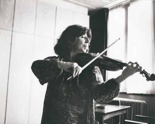 A person plays the violin in a softly lit room. The individual appears focused, wearing a patterned, long-sleeve outfit. Sunlight filters through a window with sheer curtains, creating a serene atmosphere.