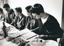 A group of people sitting around a table, likely engaged in a meal. One person, who is wearing a kippah, is reading from a book. The table is set with plates, a bottle of wine, and various dishes of food. The image is in black and white.