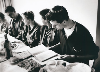 A group of people sitting around a table, likely engaged in a meal. One person, who is wearing a kippah, is reading from a book. The table is set with plates, a bottle of wine, and various dishes of food. The image is in black and white.