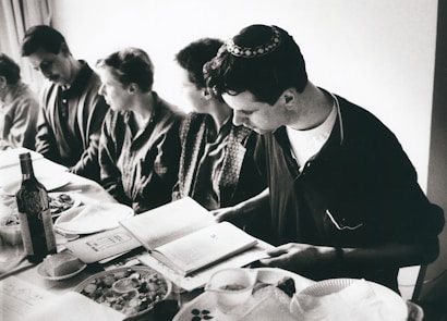 A group of people sitting around a table, likely engaged in a meal. One person, who is wearing a kippah, is reading from a book. The table is set with plates, a bottle of wine, and various dishes of food. The image is in black and white.