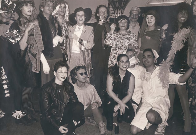A group of people dressed in a variety of costumes, including hats, dresses, and accessories, posing together in a lively manner. Some are in theatrical makeup, adding to the festive and playful atmosphere. The setting appears to be indoors, possibly at a party or event.