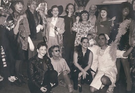 A group of people dressed in a variety of costumes, including hats, dresses, and accessories, posing together in a lively manner. Some are in theatrical makeup, adding to the festive and playful atmosphere. The setting appears to be indoors, possibly at a party or event.