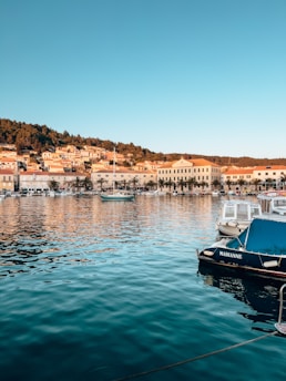 A serene waterfront scene features small boats docked at a marina. In the background, a hillside is dotted with charming buildings, reflecting Mediterranean architecture, surrounded by lush greenery. The calm water beautifully mirrors the town's buildings and skies, creating a picturesque and tranquil setting.