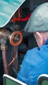 A person wearing a blue jacket and a checkered cap is operating or inspecting a piece of machinery. The machinery features a prominent orange valve with engraved text, copper pipes, and various metallic components.