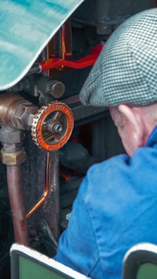 A person wearing a blue jacket and a checkered cap is operating or inspecting a piece of machinery. The machinery features a prominent orange valve with engraved text, copper pipes, and various metallic components.