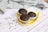 Three chocolate muffins with chunks of chocolate are placed on a yellow leaf-shaped plate. The plate sits on a white wooden surface, accompanied by scattered chocolate chips. A textured gray cloth is casually draped in the background.