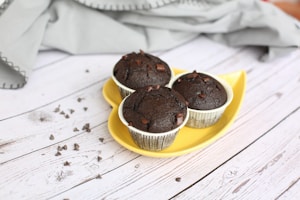 Three chocolate muffins with chunks of chocolate are placed on a yellow leaf-shaped plate. The plate sits on a white wooden surface, accompanied by scattered chocolate chips. A textured gray cloth is casually draped in the background.