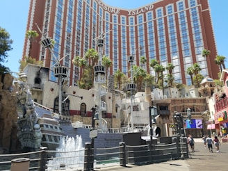A large hotel building with the signage 'Treasure Island' stands prominently in the background. In the foreground, a pirate-themed display features a replica of a pirate ship with masts and sails, surrounded by water fountains. There are palm trees scattered around the area, enhancing the tropical atmosphere. People are walking along a boardwalk, adding a sense of activity and liveliness to the scene.