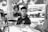 A black and white photo of a man with short hair and a beard wearing a polo shirt. He is behind a counter with various bottles and a coffee machine, looking to the side. The setting appears to be a cafe or bar with blurred patrons and tables in the background.