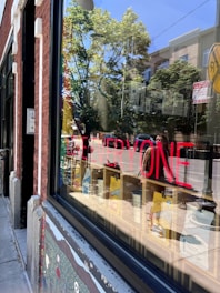 A storefront window reflects trees and buildings in a sunny outdoor setting. Bright red text on the window reads 'LOVE EVERYONE'. Inside, there are colorful objects on shelves, and outside, there's a sidewalk with a mural painted on the building's facade.