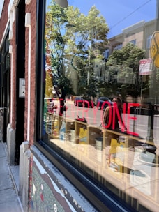 A storefront window reflects trees and buildings in a sunny outdoor setting. Bright red text on the window reads 'LOVE EVERYONE'. Inside, there are colorful objects on shelves, and outside, there's a sidewalk with a mural painted on the building's facade.