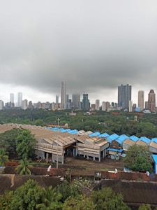 An industrial area with several warehouses featuring blue and brown roofs, set against a backdrop of high-rise buildings and lush greenery under a cloudy sky.