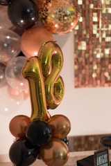 A decorative arrangement featuring golden number balloons '18' surrounded by clusters of balloons in shades of black, gold, and clear. In the background, there is a mosaic-like wall design with various shades of brown and gold. The setting suggests a celebratory event, possibly an 18th birthday party.