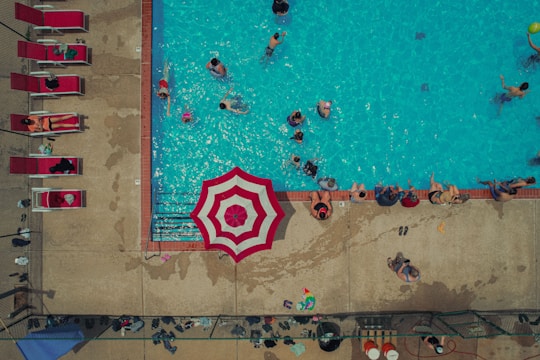 An aerial view of a swimming pool with numerous people enjoying the water. There are additional people lounging on red sunbeds beside the pool. A prominent red and white striped umbrella near the pool's edge provides some shade. The pool area is surrounded by a concrete deck that is wet from splashes of water.