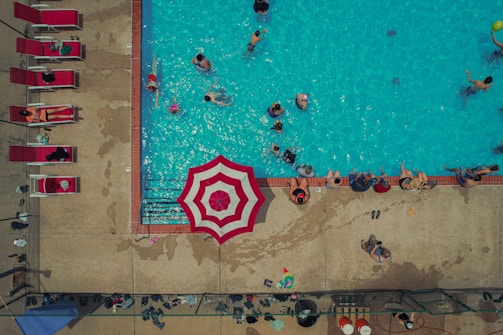 An aerial view of a swimming pool with numerous people enjoying the water. There are additional people lounging on red sunbeds beside the pool. A prominent red and white striped umbrella near the pool's edge provides some shade. The pool area is surrounded by a concrete deck that is wet from splashes of water.