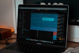 A laptop displaying digital audio workstation software with a project open on the screen, showing various audio tracks in a timeline format. The software interface has multiple panels with track names and audio waveforms visible. The room is softly lit, as indicated by the warm glow on the left side.