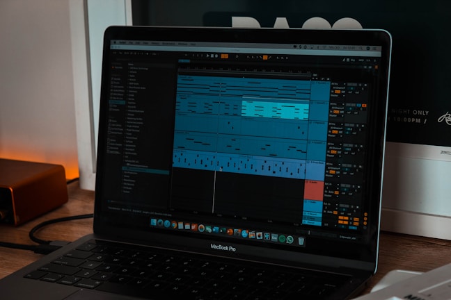 A laptop displaying digital audio workstation software with a project open on the screen, showing various audio tracks in a timeline format. The software interface has multiple panels with track names and audio waveforms visible. The room is softly lit, as indicated by the warm glow on the left side.
