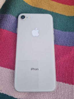 A smartphone with a white back and a single camera is placed on a colorful striped fabric. The back of the phone displays the Apple logo and the word 'iPhone' printed below.