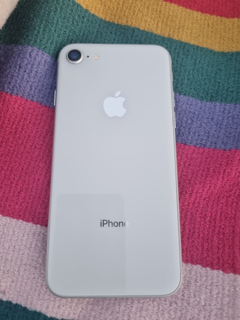 A smartphone with a white back and a single camera is placed on a colorful striped fabric. The back of the phone displays the Apple logo and the word 'iPhone' printed below.